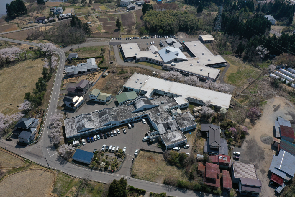 複数棟からなる介護施設の空撮。白いルーフの大型施設と駐車場が見える。