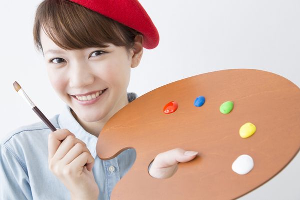 絵筆とパレットを持って「デザインは任せて」と言ってほほ笑むイメージの若い女性の画像